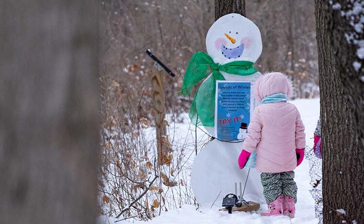 Child standing in front of a snowman sign on a snowy trail at Frosty Fest at Red Oak Nature Center.